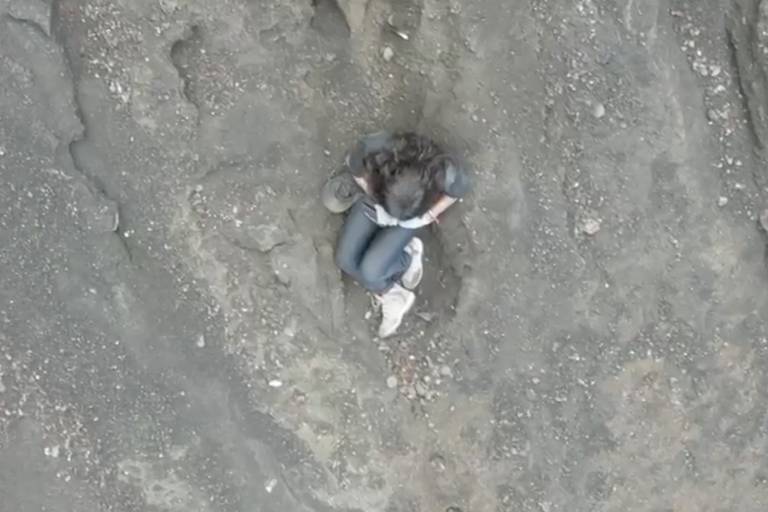 L'immagine mostra una persona seduta su una superficie rocciosa e irregolare, con il corpo curvo e le gambe piegate. La persona indossa una maglietta e jeans e sembra essere in posizione riflessiva o di riposo. Il terreno circostante è composto da rocce e terra, con alcune piccole pietre visibili.