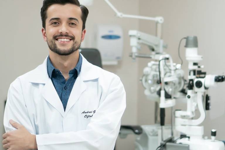 Um homem jovem, vestido com um jaleco branco, está sorrindo e posando com os braços cruzados em um consultório de oftalmologia. Ao fundo, há um equipamento oftalmológico, incluindo um oftalmoscópio e uma cadeira de exame. O ambiente é bem iluminado e organizado.
