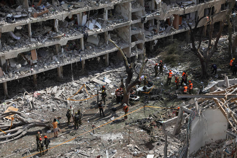 A imagem mostra os destroços de um edifício parcialmente destruído, com escombros espalhados pelo chão. Várias pessoas, vestindo coletes laranjas, estão trabalhando na área, possivelmente em atividades de resgate ou busca. Árvores danificadas estão visíveis ao redor, e há uma faixa de segurança delimitando a área. O cenário é de destruição e atividade de emergência.