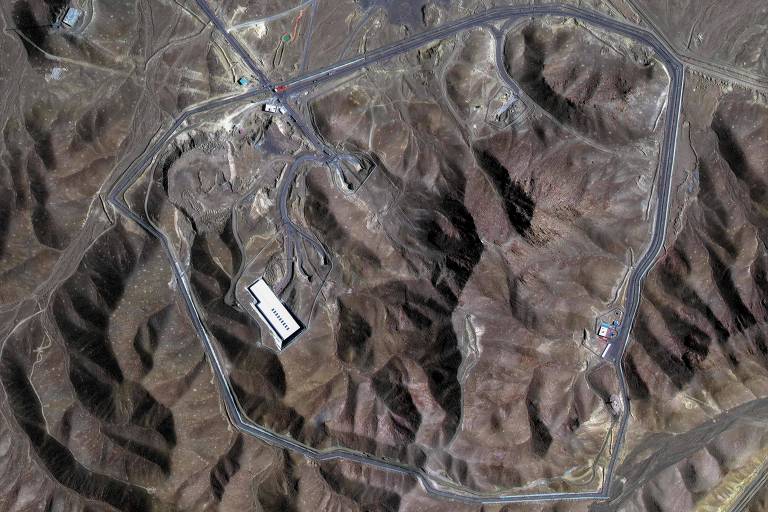 A imagem mostra uma vista aérea de uma área montanhosa com várias estradas e um grande edifício retangular branco localizado em uma das áreas. O terreno é predominantemente árido e acidentado, com várias elevações e depressões visíveis. Estradas se entrelaçam na paisagem, conectando diferentes pontos da região.