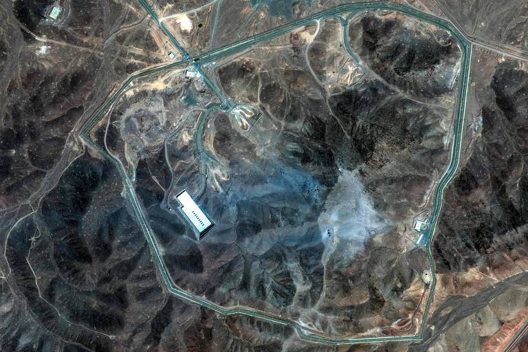 A imagem mostra uma vista aérea de uma instalação em uma área montanhosa. A estrutura principal é um edifício retangular branco localizado na parte inferior da imagem. Estradas se entrelaçam ao redor da instalação, formando um contorno que parece ser uma cerca ou limite. O terreno é árido e montanhoso, com várias formações rochosas visíveis ao redor.