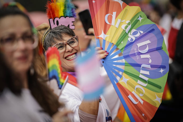 A imagem mostra uma mulher sorridente segurando um leque colorido com as palavras 'drag brunch' em destaque. Ela usa um acessório de cabeça com a palavra 'LOVE' e óculos. Ao fundo, outras pessoas também seguram bandeiras coloridas, criando um ambiente festivo e alegre, típico de celebrações do orgulho LGBTQ+. O cenário é vibrante e cheio de cores.