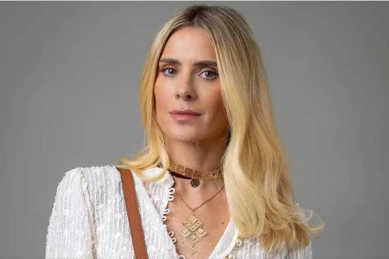 A imagem mostra uma mulher com cabelo loiro e liso, usando uma blusa branca com detalhes texturizados. Ela está com um olhar sério e levemente de lado, e usa colares e um choker. A mulher também tem uma bolsa marrom ao ombro. O fundo da imagem é cinza claro, destacando a figura da mulher.
