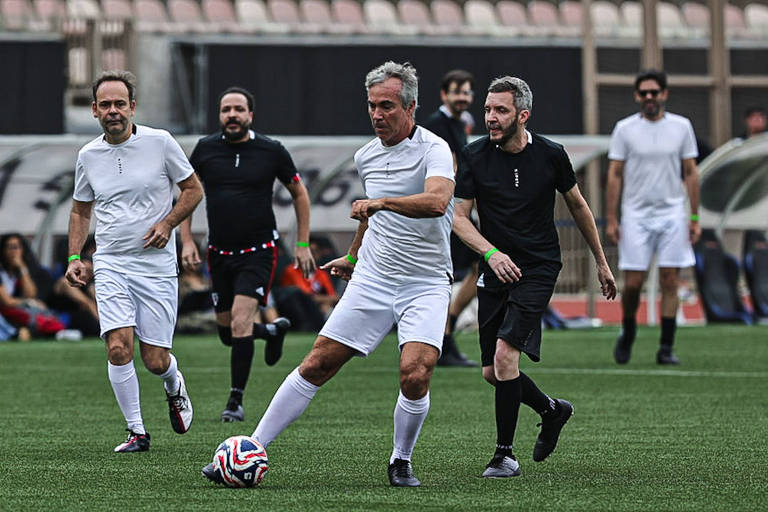 A imagem mostra um grupo de homens jogando futebol em um campo. Eles estão vestidos com uniformes de futebol, alguns em camisetas brancas e outros em camisetas pretas. Um dos jogadores, que está em destaque, está prestes a chutar a bola, que é colorida. O campo é coberto de grama e há espectadores ao fundo, além de um banco de reservas.
