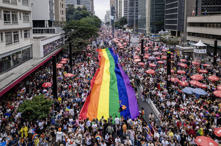 A imagem mostra uma grande multidão participando de uma parada do orgulho LGBTQIA+, com uma enorme bandeira do arco-íris sendo carregada. O evento ocorre em uma rua movimentada, cercada por prédios altos. Há muitas pessoas, algumas usando roupas coloridas e acessórios festivos. O clima é de celebração e diversidade, com barracas e mesas ao fundo, possivelmente vendendo alimentos e bebidas.