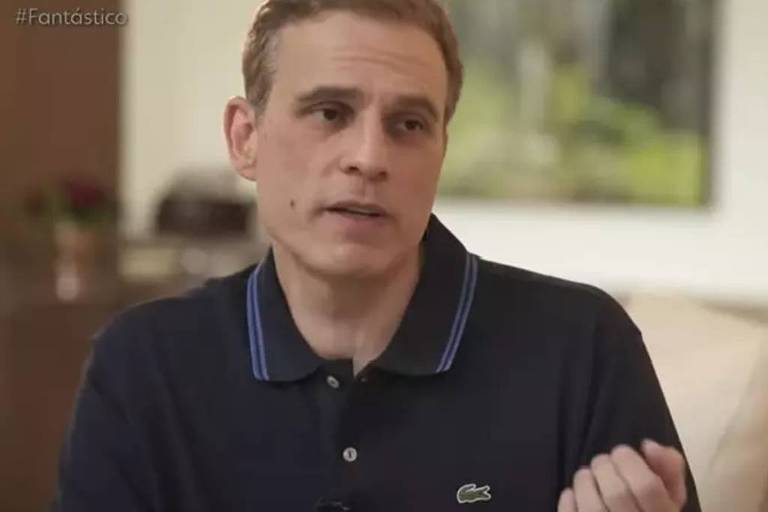 Um homem está sentado em um ambiente interno, aparentemente em uma entrevista. Ele tem cabelo curto e usa uma camiseta polo escura com detalhes em azul. O fundo é desfocado, mas parece haver uma área de estar com uma planta e uma parede clara. O homem está gesticulando com a mão direita enquanto fala.
