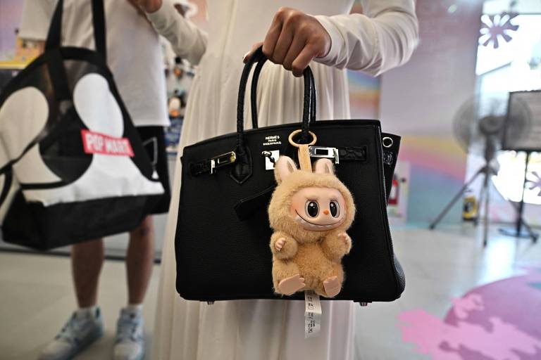 A imagem mostra uma pessoa segurando uma bolsa preta com alças. Na bolsa, há um chaveiro de pelúcia em forma de um personagem fofinho, com orelhas e um rosto sorridente. Ao fundo, é possível ver um ambiente colorido e decorado, com uma bolsa de tecido em preto e branco ao lado da pessoa.