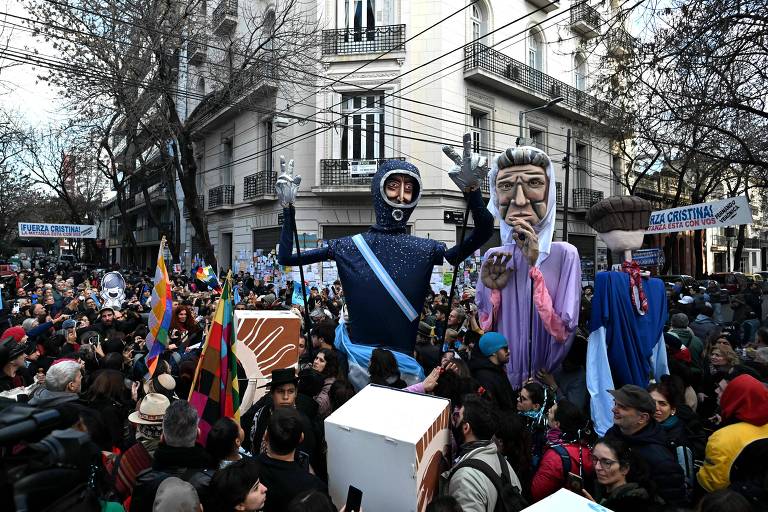 A imagem mostra uma grande manifestação com uma multidão reunida em uma rua. No centro, destacam-se dois bonecos gigantes: um homem vestido de azul e um outro com uma roupa rosa, ambos com expressões faciais marcantes. Ao fundo, há um edifício e várias pessoas segurando bandeiras coloridas. A atmosfera é de protesto ou celebração, com cartazes visíveis.