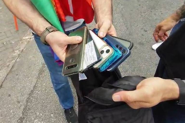 L'immagine mostra due persone per strada, una con diversi cellulari in una mano e l'altra con una borsa nera nell'altra. I cellulari sono di colori e modelli diversi. L'ambiente è urbano, con un marciapiede visibile sullo sfondo.