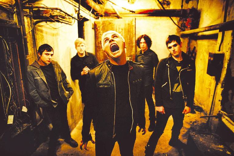 A imagem mostra cinco membros de uma banda em um ambiente escuro e industrial. O vocalista, em destaque, está com a boca aberta, como se estivesse cantando ou gritando. Os outros membros estão posicionados ao fundo, com expressões sérias. Todos estão vestidos com roupas escuras, e o ambiente é iluminado por uma luz amarelada, destacando as paredes desgastadas e os fios expostos.