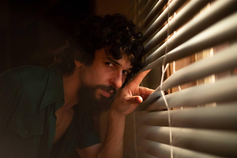 Um homem com cabelo encaracolado e barba está espiando por entre as persianas de uma janela. Ele tem uma expressão intensa e curiosa, com um olhar focado. A iluminação é suave, criando um ambiente misterioso. O homem usa uma camisa de cor escura e está em um ambiente interno.