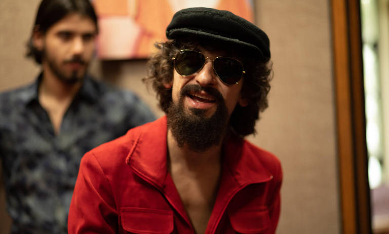 A imagem mostra dois homens em um ambiente interno. O homem à frente usa óculos escuros, um chapéu preto e uma jaqueta vermelha. Ele tem uma barba e cabelo encaracolado. O homem ao fundo está parcialmente visível, vestindo uma camisa azul com padrões. O ambiente parece ser um estúdio ou sala com paredes de tom neutro.