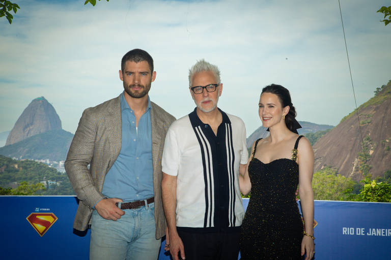 Três pessoas posam para a foto em um evento ao ar livre no Rio de Janeiro. Ao fundo, é possível ver montanhas e uma vegetação exuberante. A primeira pessoa, à esquerda, usa uma camisa clara e um blazer. A segunda pessoa, no centro, usa uma camisa branca com detalhes pretos e óculos. A terceira pessoa, à direita, usa um vestido preto e sorri. A imagem tem uma faixa azul na parte inferior com o texto 'RIO DE JANEIRO'.