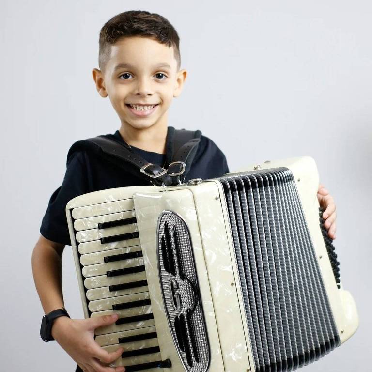 Um menino sorridente está segurando uma sanfona. Ele tem cabelo curto e usa uma camiseta preta. O fundo da imagem é cinza claro, destacando o menino e o instrumento musical que ele está segurando.