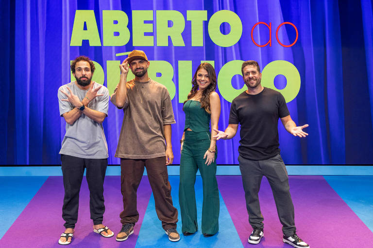 Quatro pessoas estão em um estúdio de gravação, posando para a foto. Ao fundo, há um grande painel com a inscrição 'ABERTO ao PÚBLICO'. As pessoas estão vestidas de maneira casual: um homem à esquerda com cabelo encaracolado e barba, usando uma camiseta cinza e calças escuras; um homem no centro, com um chapéu e camiseta marrom, calças escuras; uma mulher à direita, vestindo um macacão verde; e um homem à direita, usando uma camiseta preta e calças escuras. Todos estão sorrindo e fazendo gestos amigáveis.