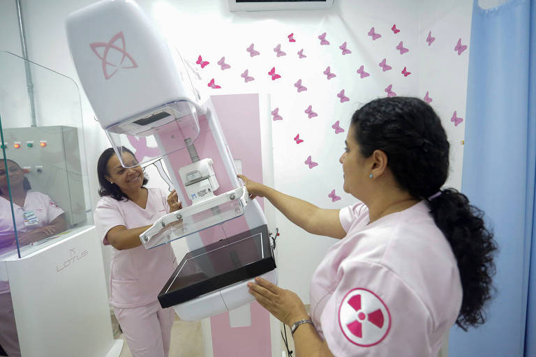 A imagem mostra duas profissionais de saúde em um ambiente clínico. Uma delas está operando um equipamento de mamografia, que é predominantemente branco com detalhes em rosa. O fundo da sala é decorado com borboletas rosa, e há um painel com luzes e botões visíveis. As profissionais estão vestidas com uniformes rosa claro.