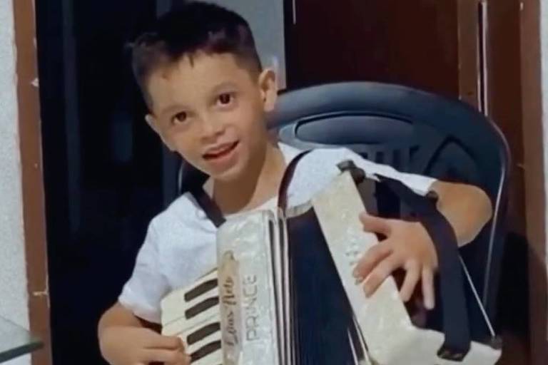 Um menino pequeno está sentado em uma cadeira plástica preta, tocando uma sanfona. Ele está sorrindo e usando uma camiseta branca. O ambiente ao fundo parece ser uma casa, com uma porta e uma parede visíveis.