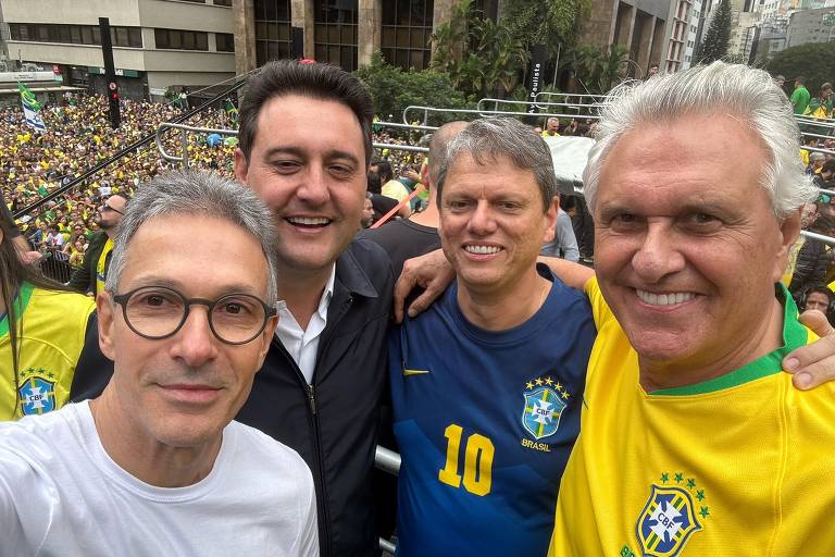Quatro homens posam para uma selfie em um evento ao ar livre, com uma grande multidão ao fundo. Eles estão sorrindo e vestindo camisetas, sendo que um deles usa uma camiseta amarela com detalhes verdes, representando as cores da bandeira do Brasil. O ambiente é festivo, com pessoas visíveis na multidão, algumas vestindo roupas nas cores da bandeira brasileira.