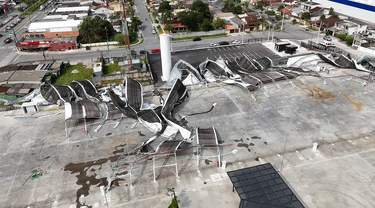A imagem mostra uma área externa com uma estrutura de cobertura danificada, onde as lonas estão rasgadas e desordenadas. O chão está molhado, possivelmente devido à chuva. Ao fundo, há uma rua com casas e estabelecimentos comerciais visíveis. A estrutura parece ter sido afetada por ventos fortes ou uma tempestade, resultando em danos significativos.