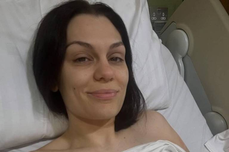 Uma mulher está deitada em uma cama de hospital, com um lençol cobrindo parcialmente seu corpo. Ela tem cabelo escuro e liso, e está sorrindo para a câmera. A imagem mostra um ambiente hospitalar, com equipamentos médicos visíveis ao fundo.