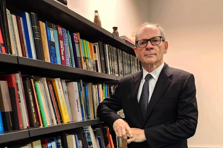 Een man in een zwart pak met stropdas en een bril poseert naast een boekenkast vol boeken. Hij heeft zijn armen over elkaar en een lichte glimlach op zijn gezicht. De kamer is goed verlicht, met een lichte muur en een blauwe vloer. Op de achtergrond staan een bureau en nog meer boeken op de plank.