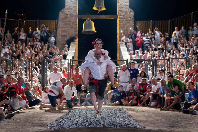 A imagem mostra um evento cultural ao ar livre, onde um homem está realizando uma performance no centro de uma arena. Ele está vestido com uma camisa branca e calças escuras, carregando uma mulher em seus braços. Ao fundo, há uma grande multidão assistindo, com pessoas de diferentes idades e vestimentas. O ambiente é iluminado por luzes e há duas grandes sinos visíveis acima da cena. O chão da arena é coberto por pedras pequenas.