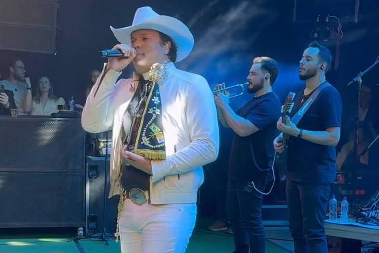 Um artista está se apresentando em um palco, segurando um microfone. Ele usa um traje branco, incluindo um chapéu e botas. Ao fundo, há dois músicos tocando instrumentos, um deles com uma guitarra. O ambiente é iluminado e há uma plateia visível ao fundo.