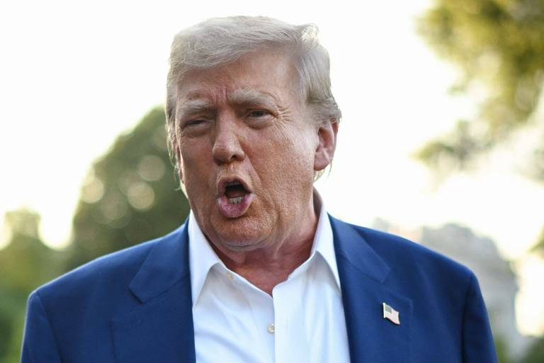 Um homem com cabelo loiro e uma expressão facial intensa está falando ao ar livre. Ele usa um paletó azul e uma camisa branca. Ao fundo, há árvores e uma luz suave, sugerindo que é um dia ensolarado.