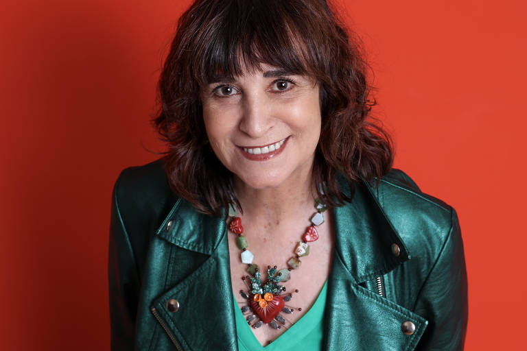 A imagem mostra uma mulher sorridente com cabelo castanho e ondulado, vestindo uma jaqueta verde e uma blusa verde por baixo. Ela usa um colar colorido com um pingente em forma de coração. O fundo da imagem é laranja, criando um contraste vibrante com a roupa da mulher.