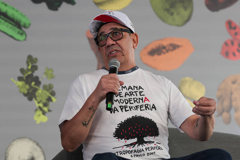 Um homem com óculos e boné, vestindo uma camiseta branca com uma estampa que diz 'Semana de Arte Moderna na Periferia' e uma ilustração de uma árvore. Ele está segurando um microfone e falando, sentado em um sofá. Ao fundo, há um padrão de folhas e frutas em cores variadas.