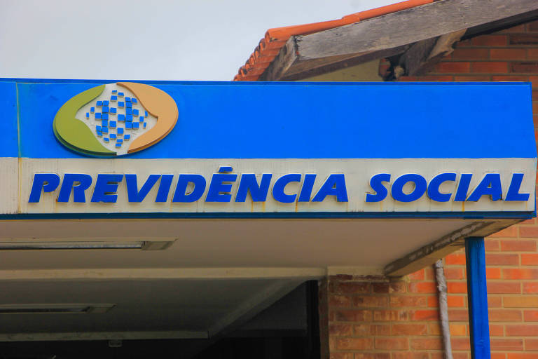A imagem mostra uma placa azul com o texto 'PREVIDÊNCIA SOCIAL' em letras brancas. Acima do texto, há um logotipo colorido que combina azul, verde e laranja. O fundo da placa é de um tom azul claro, e a estrutura ao fundo parece ser um prédio com tijolos expostos.