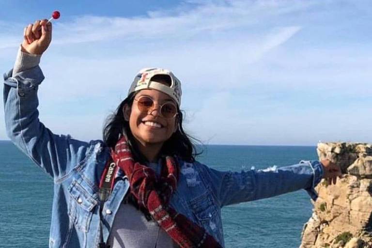 Uma pessoa sorridente está em pé em uma rocha, com o mar ao fundo. Ela usa uma jaqueta jeans, um boné e óculos de sol. A pessoa segura um doce vermelho na mão e está com os braços abertos, demonstrando alegria. O céu está claro e há algumas nuvens.