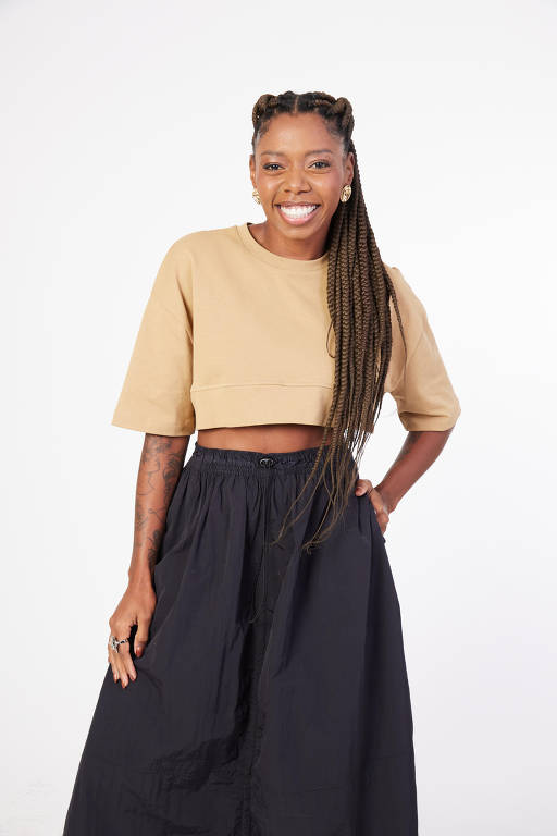 A imagem mostra uma mulher sorridente em pé, vestindo uma blusa cropped de cor bege e uma saia longa preta. Ela tem cabelo trançado e está posando com uma mão na cintura, em um fundo branco.