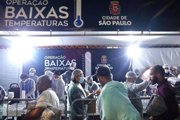 A imagem mostra um evento chamado 'Operação Baixas Temperaturas', realizado na cidade de São Paulo. Há uma tenda iluminada onde várias pessoas estão distribuindo alimentos e cobertores. Os participantes estão usando máscaras e toucas, indicando medidas de saúde e segurança. Ao fundo, há um banner com o nome da operação e o brasão da cidade de São Paulo.