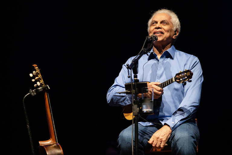 Um homem de cabelo grisalho, vestido com uma camisa azul e jeans, está sentado em um palco. Ele sorri enquanto segura um pequeno instrumento musical, possivelmente um ukulele. Ao seu lado, há um violão apoiado no chão. O fundo é escuro, destacando o artista e seu instrumento.