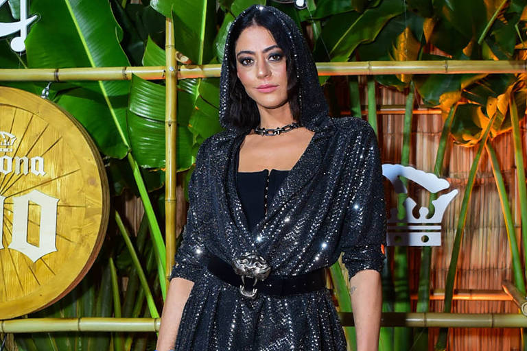 Uma mulher está posando em um evento, vestindo um traje preto brilhante com detalhes prateados. Ela usa um cinto na cintura e segura uma bolsa rosa. O fundo apresenta elementos decorativos como folhas verdes e um grande símbolo da marca Corona, além de um círculo dourado com a inscrição 'Corona 100'. O ambiente parece ser festivo e bem iluminado.