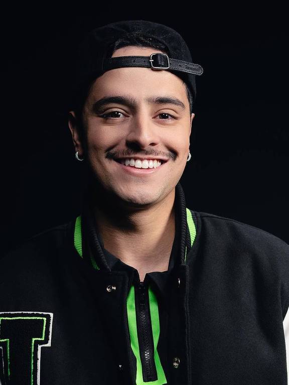 Um jovem homem está posando para a foto, sorrindo. Ele usa um boné preto e uma jaqueta estilo varsity, que é preta com detalhes em verde e branco. O fundo da imagem é escuro, destacando o rosto e a roupa do jovem.