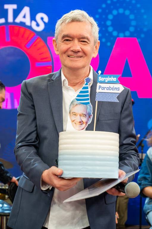 Um homem de cabelos grisalhos está segurando um bolo de aniversário. O bolo é de cor azul claro e tem uma vela em forma de chapéu de festa. Há uma foto de um homem na parte superior do bolo, e uma faixa com os dizeres 'Serginho Parabéns'. O fundo é colorido e festivo, com elementos decorativos visíveis.