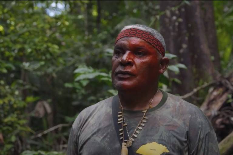 Um homem está em uma floresta densa, cercado por árvores verdes. Ele tem a pele pintada de vermelho e usa uma faixa na cabeça. Seu traje é camuflado, e ele usa um colar com um pingente. O ambiente é natural e exuberante, com folhagens ao fundo.