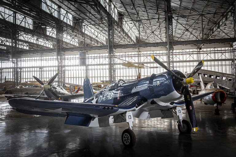 A imagem mostra um avião de caça com pintura azul escura em um hangar amplo. O ambiente é iluminado por luz natural que entra pelas janelas grandes. Ao fundo, outros aviões estão visíveis, e o chão é de concreto. O avião em destaque possui hélices e está posicionado sobre rodas.