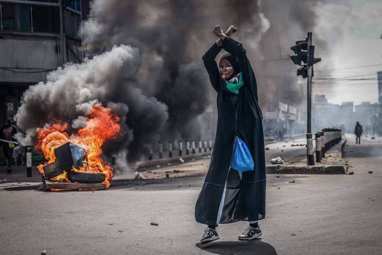 A imagem mostra uma pessoa em pé em uma rua, segurando dois bastões acima da cabeça. Ao fundo, há uma barricada em chamas feita de pneus, com fumaça densa subindo. A pessoa está vestida com uma roupa escura e uma máscara, e parece estar em um ambiente de protesto, com sinais de tumulto ao redor.