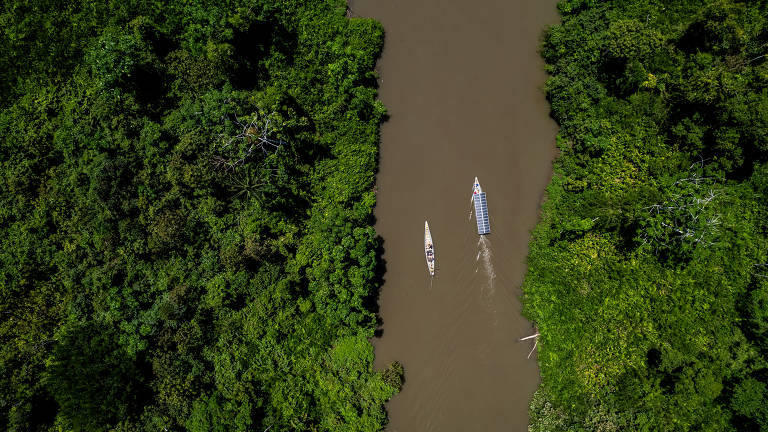 A imagem mostra uma vista aérea de um rio de água marrom cercado por densa vegetação verde da floresta. No rio, há duas canoas brancas visíveis, navegando na água. A vegetação ao redor é rica e variada, com árvores altas e folhagem abundante.