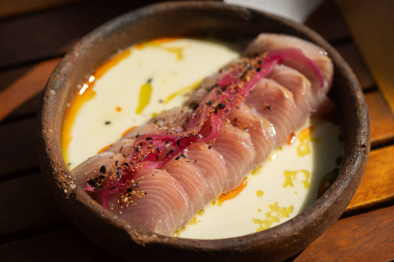 A imagem mostra um prato com fatias de peixe cru dispostas sobre um molho cremoso, decorado com cebolas roxas e temperos. Ao fundo, há uma cesta com fatias de pão. A apresentação é em um recipiente de barro e a mesa é de madeira.