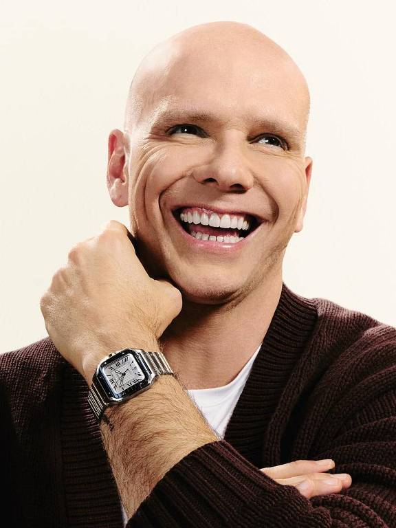 Um homem careca, sorrindo amplamente, está sentado em um sofá. Ele usa um suéter escuro e uma camiseta branca. O homem tem um relógio de pulso prateado e está com os braços cruzados, apoiando o queixo em uma das mãos. O fundo é claro e neutro, destacando sua expressão alegre.