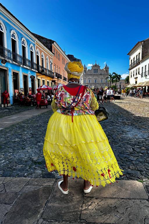 A imagem mostra uma mulher vista de costas, vestindo um traje tradicional colorido, predominantemente amarelo, com detalhes em vermelho. Ela está em uma rua de paralelepípedos, cercada por edifícios históricos de cores vibrantes. Ao fundo, há pessoas e uma igreja, sob um céu azul claro.
