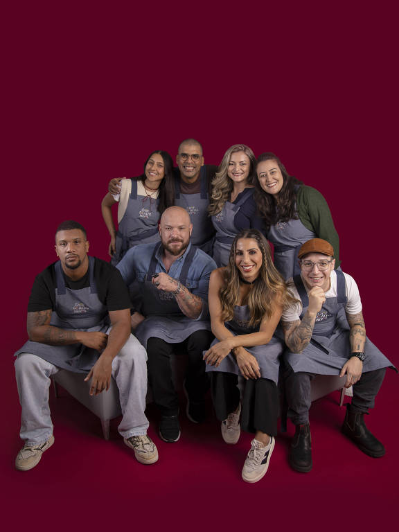 Uma foto de um grupo de oito pessoas, todas vestindo aventais, posando em um fundo vermelho. As pessoas estão organizadas em duas fileiras, com quatro na parte de trás e quatro na frente. Elas estão sorrindo e fazendo gestos amigáveis, transmitindo uma sensação de camaradagem e alegria.