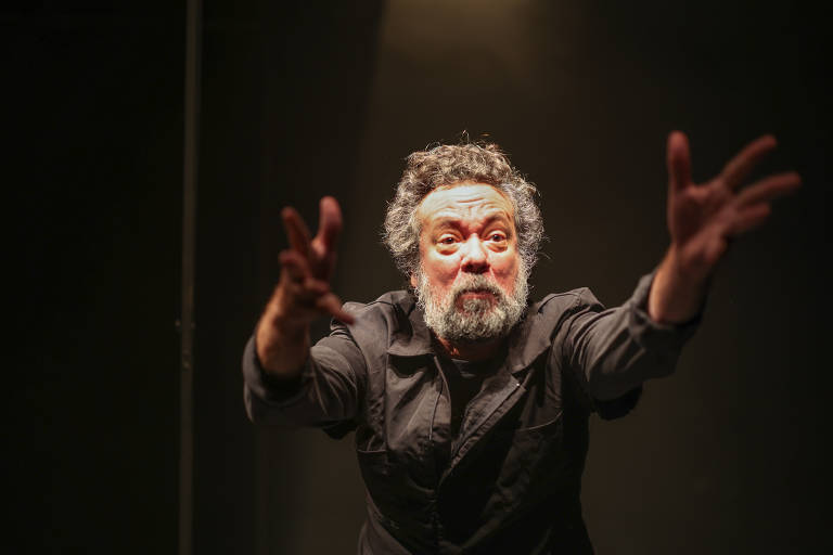 Um homem com cabelo grisalho e barba, vestindo uma roupa escura, está em uma pose expressiva, com os braços estendidos para frente. Ele parece estar em um palco, com iluminação focada em seu rosto, que demonstra emoção intensa. O fundo é escuro, destacando sua figura.