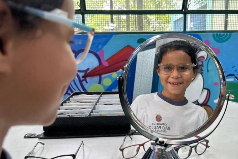A imagem mostra uma criança com óculos, sorrindo enquanto se olha em um espelho. O fundo é colorido, com grafites e desenhos. À frente da criança, há uma caixa com óculos. A iluminação é natural, e a cena parece estar em um ambiente interno.