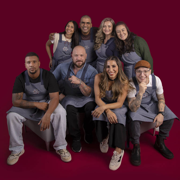 Uma foto de um grupo de oito pessoas, todas vestindo aventais, posando em um fundo vermelho. As pessoas estão organizadas em duas fileiras, com quatro na parte de trás e quatro na frente. Elas estão sorrindo e fazendo gestos amigáveis, transmitindo uma sensação de camaradagem e alegria.