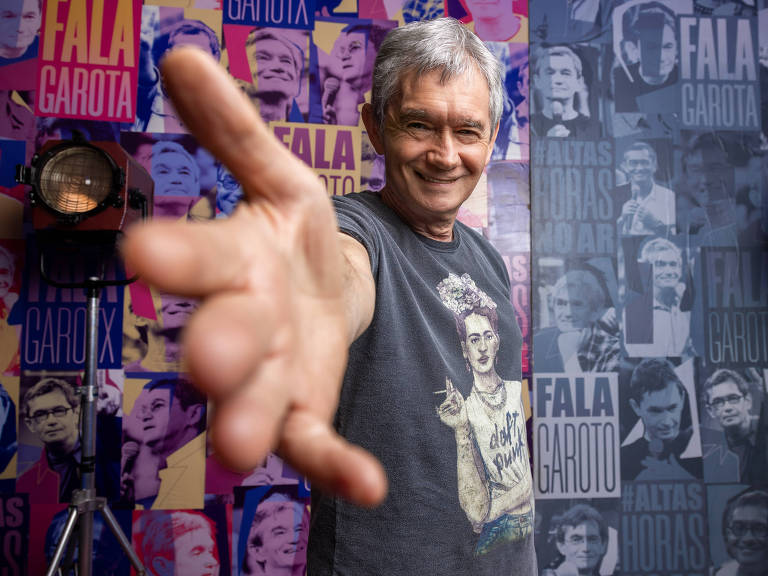 Um homem de cabelos grisalhos, usando uma camiseta preta com uma estampa de uma mulher, está posando para a foto com um gesto de saudação, estendendo a mão em direção à câmera. Ao fundo, há uma parede decorada com várias imagens e textos, incluindo as palavras 'Fala Garoto' e 'Faltas Horas'.